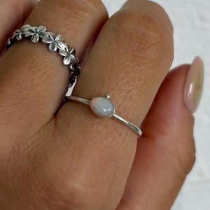 May include: A silver ring featuring a small, smooth, oval-shaped white stone set on a thin band, worn alongside a second silver ring designed with a continuous pattern of small, five-petalled flowers encircling the entire band.