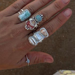 May include: Four silver rings worn on fingers. One features a rectangular blue stone, another a round blue stone above a red and white shell, a third has three shell segments, and the fourth is a thin band with a small, oval, light blue stone.
