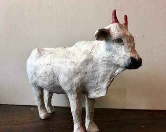 Puede incluir: Escultura de vaca blanca de papel maché con cuernos rojos. La escultura está de pie sobre una superficie de madera.