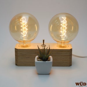 May include: A wooden table lamp featuring two large, round, amber-colored Edison bulbs. The bulbs are mounted on a layered wooden base with a small switch. A small white square pot with a succulent plant is in front.