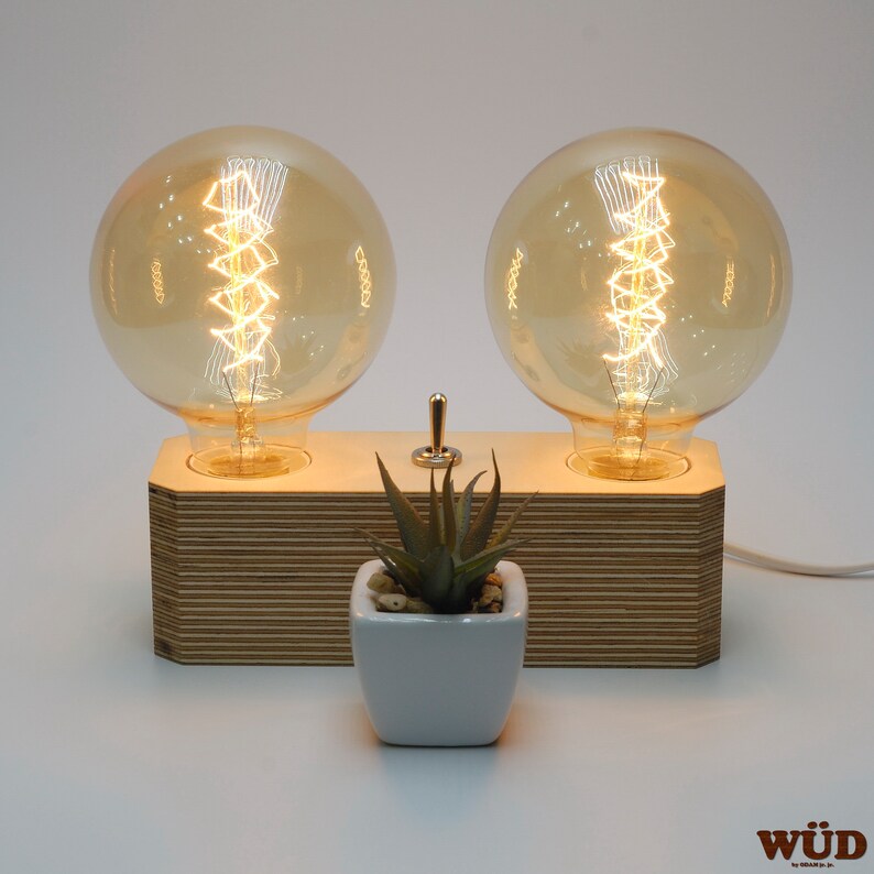 May include: A wooden table lamp featuring two large, round, amber-colored Edison bulbs. The bulbs are mounted on a layered wooden base with a small switch. A small white square pot with a succulent plant is in front.