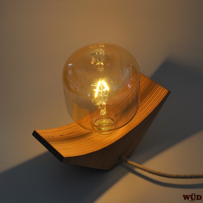 May include: A wooden wall lamp with a curved base and a clear glass light bulb. The light bulb is lit and casting a warm glow.