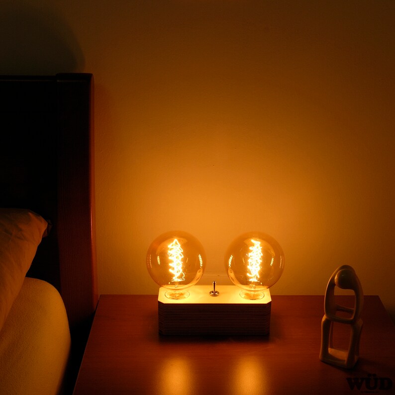 May include: A wooden bedside lamp with two large, round, Edison-style light bulbs. The bulbs are lit, emitting a warm, orange glow. The lamp has a rectangular base with a switch. A wooden sculpture is on the right.
