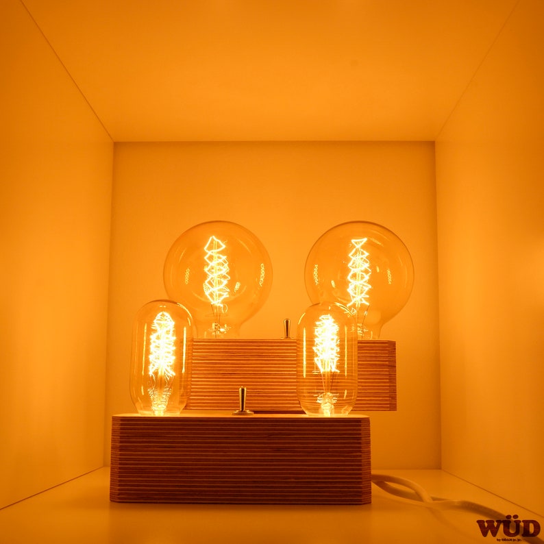May include: Four illuminated Edison-style light bulbs, two large and two smaller, are displayed on wooden platforms. The bulbs emit a warm, orange glow, creating a cozy ambiance. The arrangement is set against a white background, with a visible power cord.
