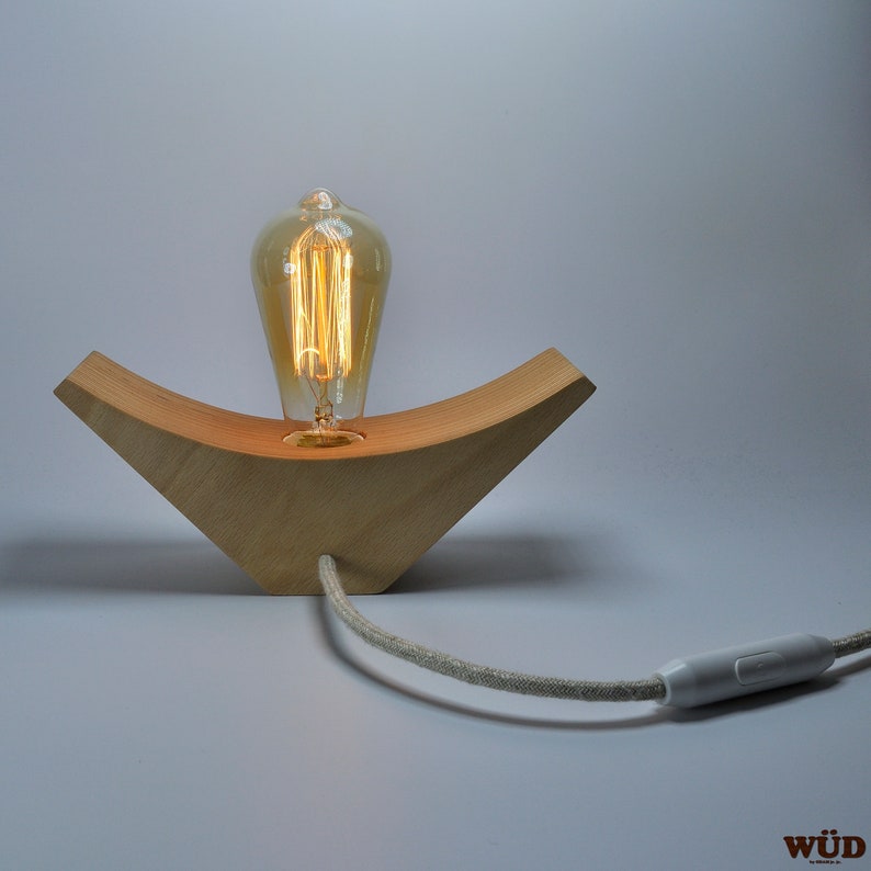 May include: A wooden lamp base with a curved design holds a vintage-style light bulb. The lamp has a linen cord with a white switch.