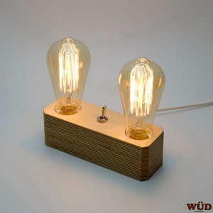 May include: A wooden table lamp with two Edison-style light bulbs. The rectangular base is made of layered wood, and a chrome toggle switch is centered between the bulbs. A white cord extends from the back.