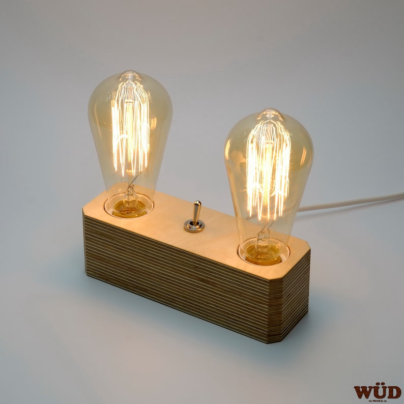 May include: A wooden table lamp with two Edison-style light bulbs. The rectangular base is made of layered wood, and a chrome toggle switch is centered between the bulbs. A white cord extends from the back.