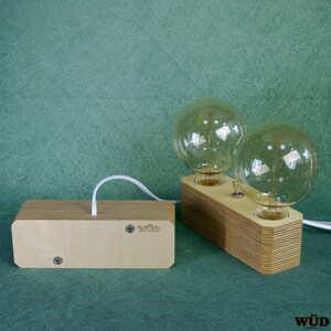 May include: A wooden desk lamp with two large, round light bulbs. The lamp has a light wood finish and a switch to turn the lights on and off. The lamp is connected to a wooden box with the word "w&uuml;d" printed on it.