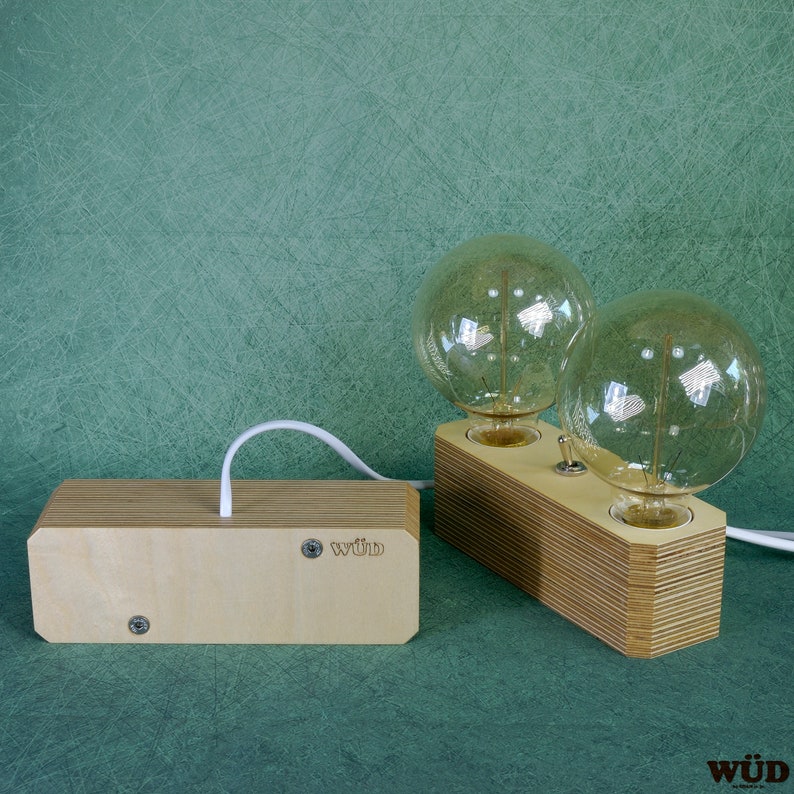 May include: A wooden desk lamp with two large, round light bulbs. The lamp has a light wood finish and a switch to turn the lights on and off. The lamp is connected to a wooden box with the word "w&uuml;d" printed on it.