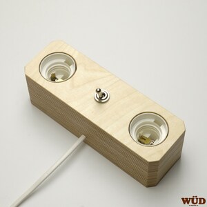May include: A light-colored wooden light fixture with two bulb sockets and a toggle switch. The fixture has a rectangular shape with a layered design and a white power cord extending from the side.