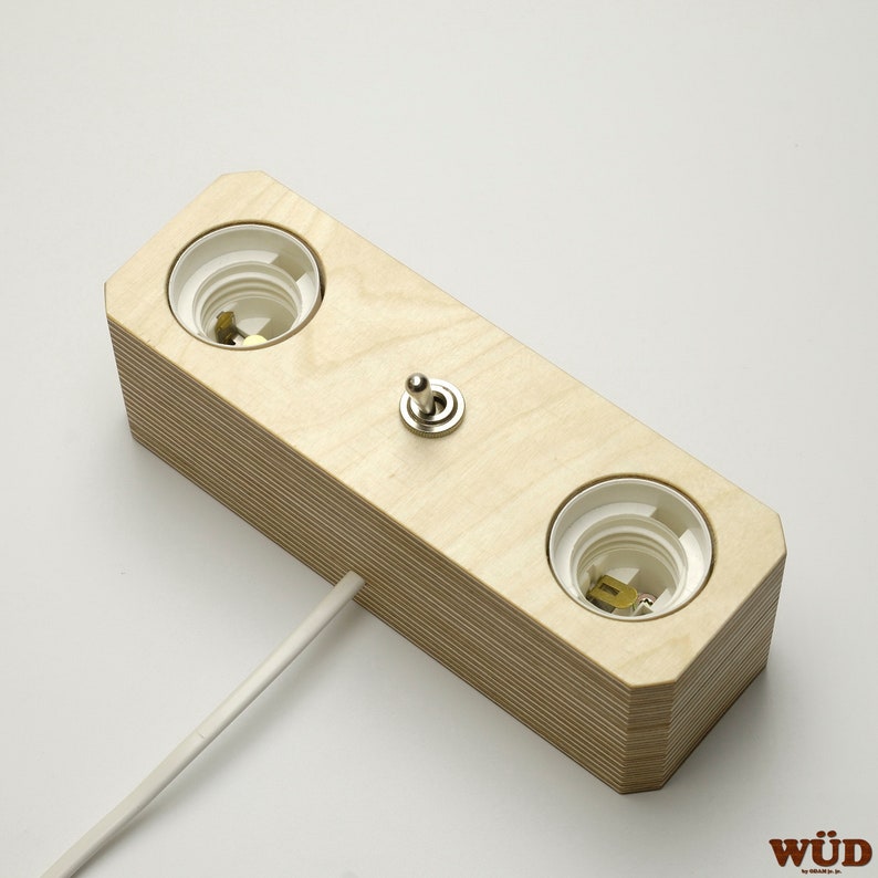 May include: A light-colored wooden light fixture with two bulb sockets and a toggle switch. The fixture has a rectangular shape with a layered design and a white power cord extending from the side.