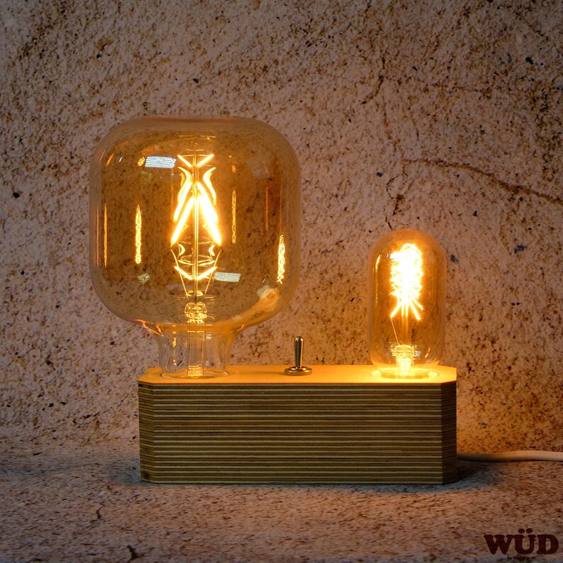 May include: A wooden desk lamp with two Edison-style light bulbs. The larger bulb is rectangular, and the smaller one is rounded. The base is made of layered wood with a switch. The bulbs emit a warm, amber glow.
