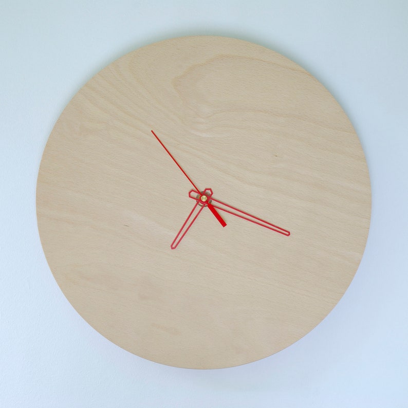 May include: A simple, round, wooden clock with red hands. The clock face is made of light wood and has a subtle grain pattern.