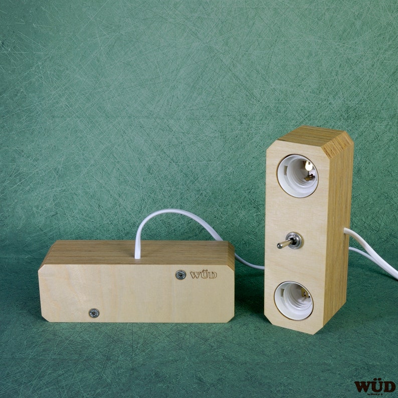 May include: A wooden lamp with two light sockets and a switch, connected to a rectangular wooden base via a white cord. The lamp and base are made of light-colored wood. The lamp has the text "w&uuml;d" on the base.