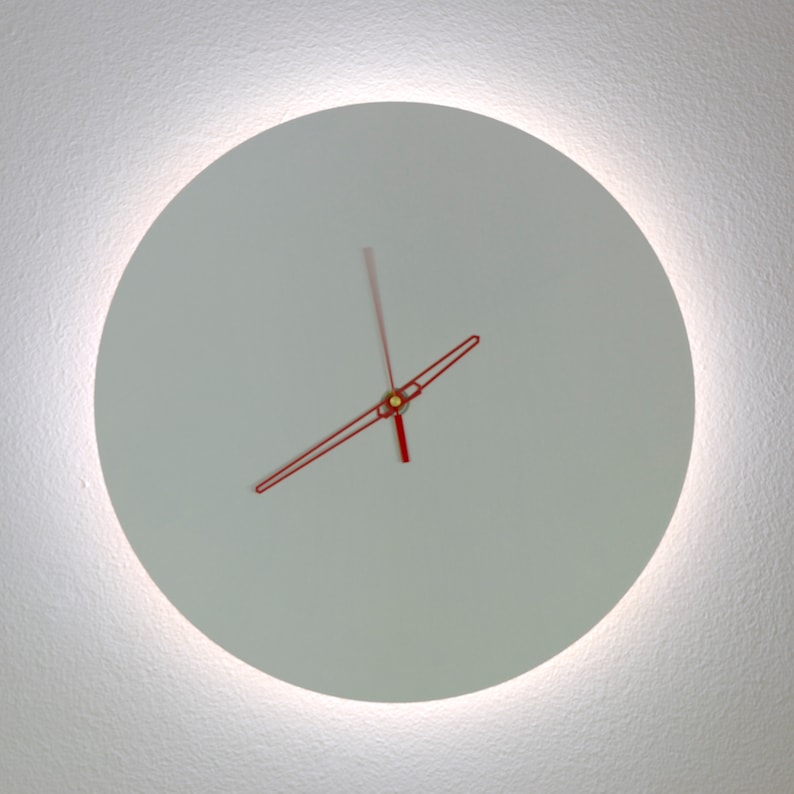 May include: A white clock face with red hands, set against a white wall. The clock is illuminated by a soft, white light.