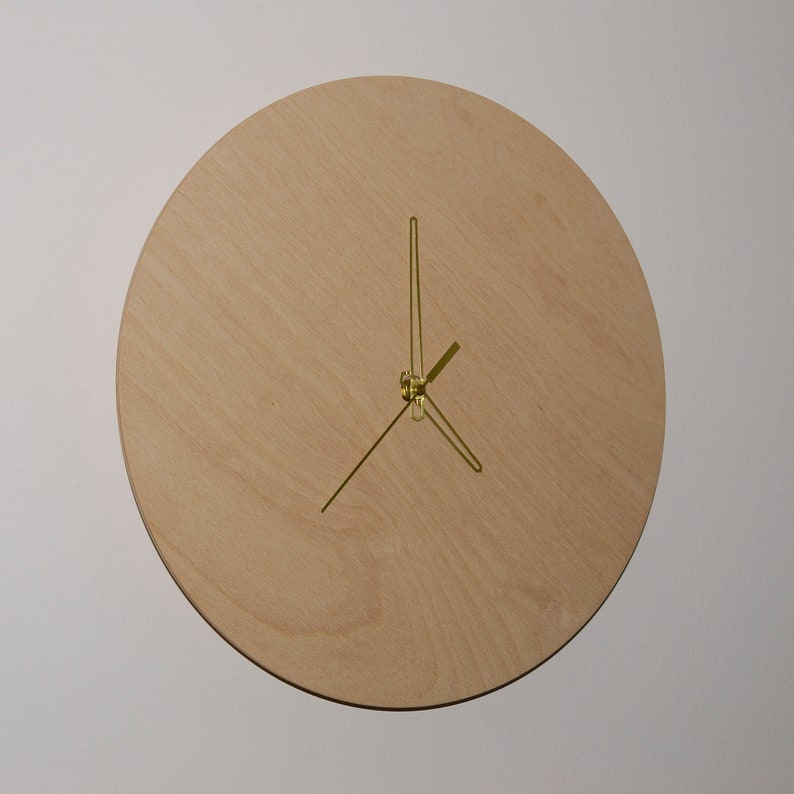 May include: A round, wooden clock with gold-colored hands. The clock face is made of light-colored wood with a subtle grain pattern.