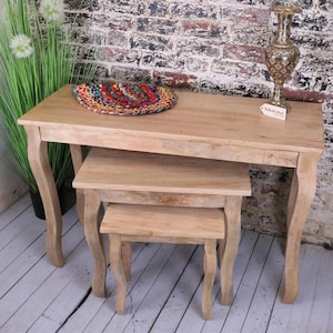 May include: A set of three wooden nesting tables with a light brown finish. The tables have curved legs and are arranged in descending size. The largest table has a colourful round rug on top.