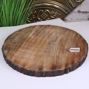 Puede incluir: Una tabla redonda de madera natural para servir con un borde de corteza rugosa. La tabla es de color marrón claro con vetas de madera visibles y una pequeña etiqueta que dice "Maravi". Adecuada para servir alimentos.