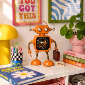 May include: Orange robot-shaped clock with a square clock face and a black background. The clock has silver accents and a small briefcase. The clock is on a white shelf with books and a plant.