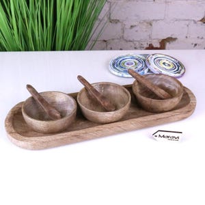 May include: A wooden serving set comprising three small bowls with matching spoons, presented on an elongated, oval tray. The bowls and spoons are crafted from light-toned wood. Ideal for serving dips, snacks, or condiments.