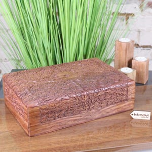 May include: A rectangular, brown wooden box with intricate floral carvings. The box is displayed on a wooden surface, with green plants and wooden candle holders in the background. The box has a detailed, textured surface.