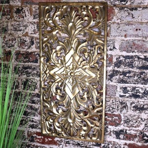 Könnte beinhalten: Goldene Wanddekoration aus Metall mit einem komplizierten, verzierten Design. Das Design zeigt eine große Diamantform mit einem floralen Muster im Inneren. Das Stück ist rechteckig und hat eine patinierte Oberfläche.