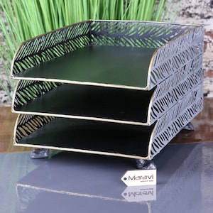 May include: A three-tier metal document tray with a black finish and gold-toned edges. The side panels feature a cut-out leaf pattern. A small rectangular tag with the text Maravi is positioned in front of the tray on the surface.