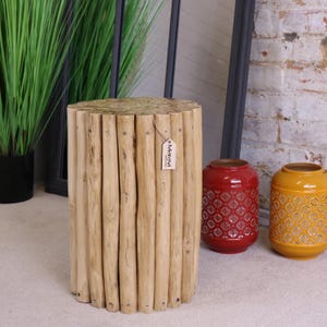 May include: A natural wood stool made of stacked logs, with a brown paper tag that reads "Maravi" on the side. The stool is sitting on a beige carpet, with two decorative lanterns in the background.