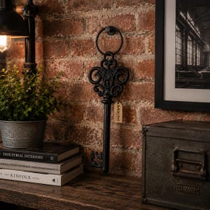 May include: A black, ornate, cast iron wall-mounted key decoration featuring a circular ring at the top and intricate scrollwork details. A small, rectangular, light-coloured paper tag with the printed text Maravi is attached to the key.