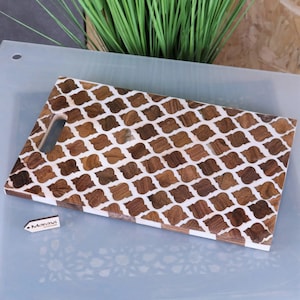 Op de afbeelding: Rechthoekig houten serveerplank met een handvat, met een herhalend patroon van bruine en witte ingelegde vormen. De plank heeft een lichtgekleurde rand en een klein label met de tekst "Maravi".