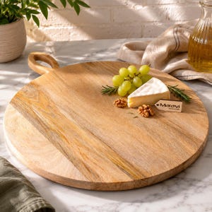 Puede incluir: Una tabla de servir redonda de madera marrón claro con asa, que presenta un racimo de uvas verdes, una cuña de queso brie y dos nueces. Una pequeña etiqueta rectangular de madera con el texto Maravi está colocada sobre la superficie.
