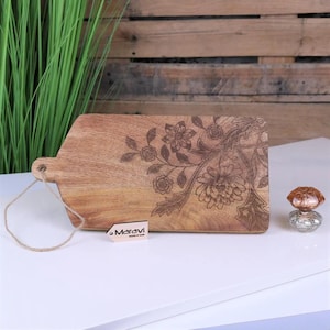 Può includere: Un tagliere in legno con un motivo floreale inciso sulla superficie. Il tagliere ha un manico e un piccolo cartellino con la scritta "Maravi" su di esso.