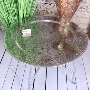 May include: A round, silver-colored metal tray table with an intricate floral pattern. The tray sits on a three-legged, silver-colored metal base. The tray has a raised edge and a decorative design. The table is suitable for indoor use.