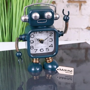 May include: A teal robot-shaped desk clock with a square clock face and a white background. The clock has a silver headset, arms, and legs. The clock face displays black numbers and hands. A small tag with the word "Maravi" is attached.