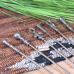 May include: Set of six fireplace tools with decorative metal handles. The tools include a poker, a shovel, a brush, a tongs, a log lifter, and a grate lifter. The tools are made of metal and have a silver finish. The tools are arranged on a woven rug with a green plant in the background.