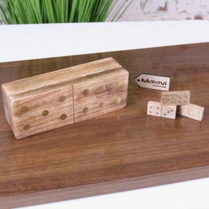 May include: Wooden domino set with a large wooden box and several individual dominoes. The box is rectangular with rounded edges and has circular indentations. The dominoes are light brown with dark brown dots. The brand name "Maravi" is on a small wooden sign.