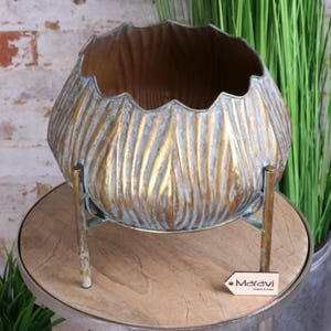 Puede incluir: Un macetero de metal dorado con un diseño texturizado en forma de hoja. El macetero está sobre un soporte con tres patas. El macetero está etiquetado con "Maravi Designed by Zara".