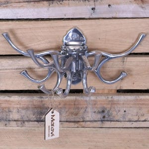 Könnte beinhalten: Silberner Metall-Kleiderhaken mit mehreren Haken. Der Haken ist an einer Holzwand montiert. Der Haken trägt ein Etikett mit der Aufschrift "Maravi Made by Hand".