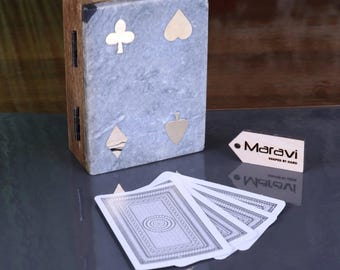 May include: A wooden playing card box with a gray marble-like front, adorned with gold-colored suit symbols. The box is next to a fanned-out deck of cards with a detailed white and gray design. The brand name "Maravi" is on a small tag.