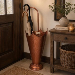 Pode incluir: Um suporte de guarda-chuva cor de cobre com textura martelada, segurando três guarda-chuvas. O suporte tem um topo em forma de coroa e uma base redonda. Os guarda-chuvas são pretos, bege e pretos. O suporte está ao lado de uma porta de madeira e de uma mesa lateral.