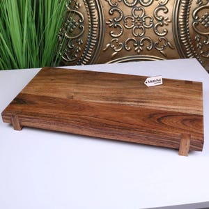 May include: A rectangular wooden cutting board with a rich brown color and a natural wood grain pattern. The board has small wooden feet and a tag that reads "Maravi". This kitchen item is ideal for food preparation.