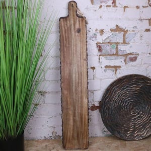 May include: A long, rectangular wooden serving board with a natural edge and a handle. The board is light brown with darker wood grain and a rough bark edge. A tag hangs from the handle. The board is displayed against a white brick wall.