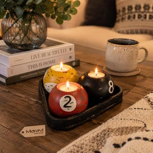May include: Three spherical candle holders designed like billiard balls in yellow, red, and black, each featuring a white circular inlay with a number. The red ball displays the number 2, the black ball shows the number 8, and all contain lit tea lights.