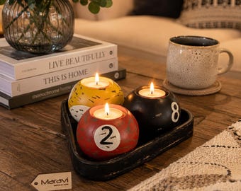 May include: Three spherical candle holders designed like billiard balls in yellow, red, and black, each featuring a white circular inlay with a number. The red ball displays the number 2, the black ball shows the number 8, and all contain lit tea lights.