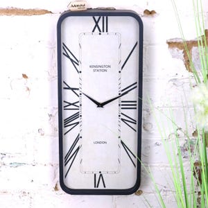 May include: Rectangular wall clock with a black frame and a white face. The clock features black Roman numerals, black hands, and the text "Kensington Station London". The clock is mounted on a white brick wall.