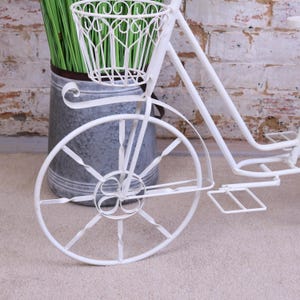 Pode incluir: Plantador de bicicleta de metal branco com uma cesta no guidão. O plantador tem um acabamento desgastado e é perfeito para exibir flores ou plantas.