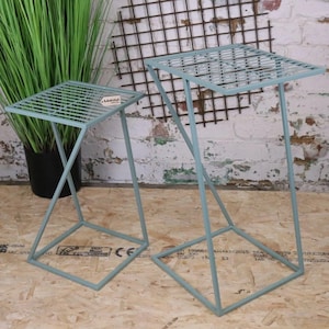 Peut inclure: Deux tables d'appoint en métal bleu clair avec un plateau carré et une base géométrique. Les plateaux ont un motif quadrillé. Les tables sont à côté d'une grande plante verte dans un pot noir. Fond de mur de briques blanches.
