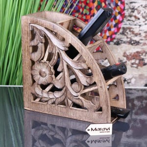 May include: A wooden remote control tidy with a floral carved design. The tidy is divided into multiple compartments and holds several black remote controls. The wood has a natural finish and sits on a reflective surface.