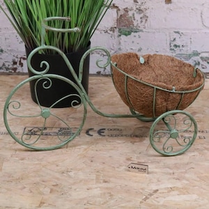 Peut inclure: Un pot de fleurs décoratif en forme de tricycle en métal vert menthe, avec un panier marron tressé. Le tricycle présente des volutes décoratives sur les roues et le guidon. Le panier est rempli d'un matériau fibreux brun, prêt à accueillir des fleurs ou des herbes.