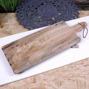 May include: A rectangular wooden cutting board with a handle. The board is light brown with visible wood grain and a natural finish. A tag with the word "Moravi" is attached. A twine loop is attached to the handle.